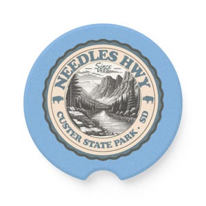May include: A light blue car coaster featuring a detailed illustration of a mountain landscape with a river, trees, and the text "Needles Hwy Since 1922 Custer State Park SD". The coaster has a scalloped edge.