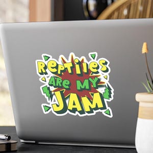 Może przedstawiać: Naklejka z napisem "Reptiles Are My Jam" w stylu komiksowym. Tekst jest żółty i zielony, obramowany na biało, z czerwono-żółtym tłem. Naklejka znajduje się na szarym laptopie.