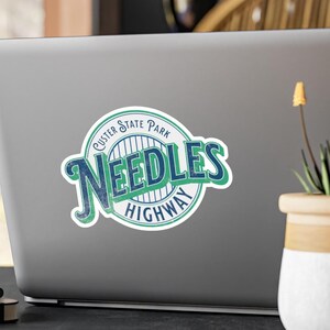 Może przedstawiać: Zielono-niebieska naklejka z tekstem "Custer State Park Needles Highway" w stylu vintage.