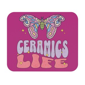 Pode incluir: Um mousepad magenta com um design retrô. Um gráfico de borboleta colorida está acima das palavras "CERAMICS LIFE" em uma fonte groovy. Acentos de estrelas amarelas pontuam o design.