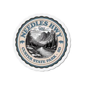 May include: A round sticker featuring a black and white illustration of a mountain landscape with a river. The sticker has a blue and tan border with the text "Needles Hwy" and "Custer State Park SD" and the year "Since 1922".