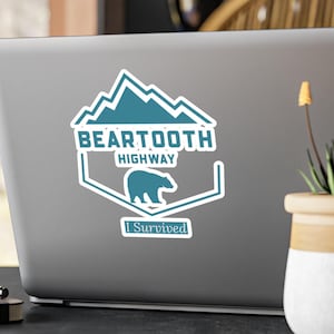 May include: A blue and white sticker with a mountain range design and the text "Beartooth Highway I Survived". A white bear silhouette is in the center of the sticker.