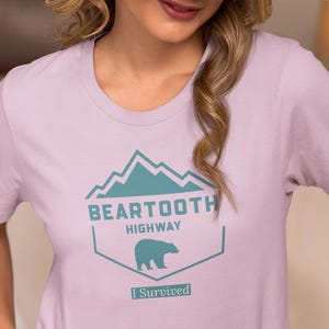 以下が含まれることがあります： 山脈とクマのターコイズブルーのグラフィックが描かれたラベンダー色のTシャツ。グラフィックには「ベアトゥースハイウェイ」と「I Survived」の文字が書かれています。