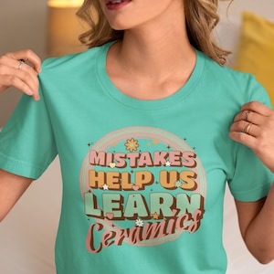 May include: A turquoise t-shirt with a retro design. The design features the text "Mistakes Help Us Learn Ceramics" in a layered, colorful font with stars and flower accents. The shirt is a crew neck style.