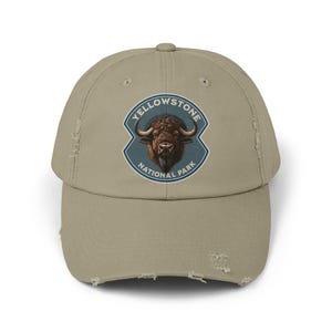 May include: Tan baseball cap with a distressed look. The cap features a circular patch with the text "YELLOWSTONE NATIONAL PARK" around a bison illustration. The cap has a curved brim and a worn appearance.
