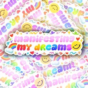May include: A sticker with the text "manifesting my dreams" in colorful bubble letters, surrounded by rainbows, hearts, and smiley faces. The background features similar designs in pastel colors.