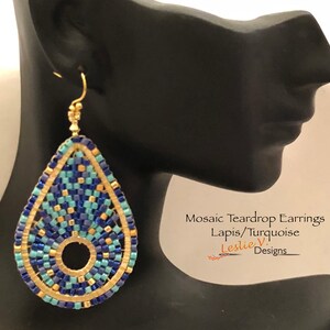 May include: Mosaic teardrop earrings in blue, gold, and turquoise colours. The earrings are made of beads and have a teardrop shape with a circular opening in the centre. The earrings are hanging from a mannequin's ear. The text "Mosaic Teardrop Earrings Lapis/Turquoise Leslie V. Designs" is visible below the earrings.