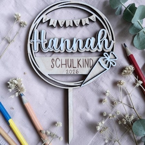 Könnte beinhalten: Ein runder Tortenstecker aus Holz mit dem Namen Hannah in erhabener weißer Schreibschrift. Über dem Namen befindet sich ein Banner mit den Buchstaben A B C D E F G. Darunter steht der Text SCHULKIND 2026 und eine Grafik einer Schultüte.