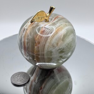 May include: A green, brown, and white marble apple with a gold leaf on top. The apple is about the size of a UK 20 pence coin.