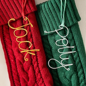 Puede incluir: Dos calcetines navideños de punto trenzado, uno rojo y otro verde, cuelgan de una guirnalda. El calcetín rojo tiene una etiqueta con el nombre "Jack" en alambre dorado, y el calcetín verde tiene una etiqueta con el nombre "Polly" en alambre plateado. Adornos y vegetación decoran los calcetines.
