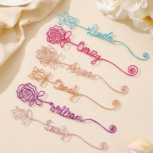 May include: A selection of wire rose bookmarks in various colours, each with a name written in cursive. The colours include blue, pink, peach, copper, purple, and lavender. The names are Linda, Crazy, Anna, Candy, William, and Emily.