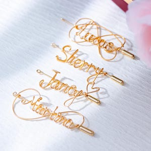 May include: Four gold-colored wire hairpins with names: Lucas, Sherry, Jancy, and Martina. Each hairpin features a unique cursive name design. The hairpins are displayed on a white surface, with a blurred pink flower and a burgundy box in the background.