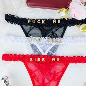 May include: Three pairs of lingerie in black, white, and red, each with a different message in gold lettering: "FUCK ME", "BAD GIRL", and "KISS ME". The lingerie features lace detailing and a thong style.