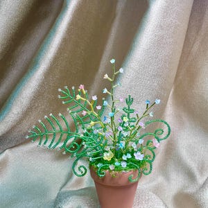 May include: A handcrafted beaded floral arrangement in a small terracotta pot. The arrangement features green beaded leaves and stems, with delicate pastel-coloured flowers. The pot sits on a shimmering, golden-toned fabric.