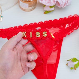 Peut inclure: String rouge avec bordure en dentelle et le mot "KISS" en lettres dor&eacute;es avec des strass. La lingerie est faite d'un mat&eacute;riau transparent.