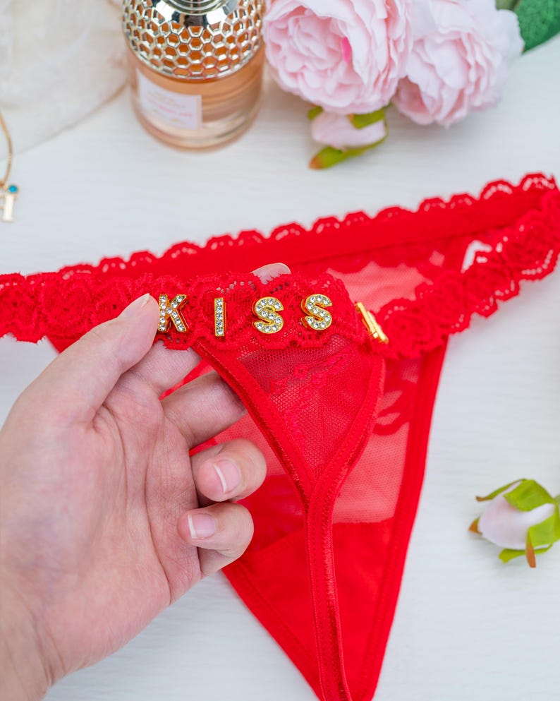 Peut inclure: String rouge avec bordure en dentelle et le mot "KISS" en lettres dor&eacute;es avec des strass. La lingerie est faite d'un mat&eacute;riau transparent.