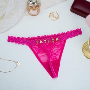 Peut inclure: String en dentelle rose vif avec le nom "TAYLOR" en lettres dor&eacute;es. Les bords sont orn&eacute;s de dentelle en forme de c&oelig;ur. Un collier en or, un flacon de parfum et d'autres accessoires sont en arri&egrave;re-plan.