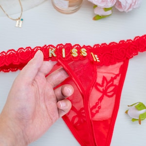 Peut inclure: String rouge avec le mot "KISS" &eacute;pel&eacute; en lettres de strass dor&eacute;es. Le sous-v&ecirc;tement a une bordure en dentelle et un motif floral sur le devant. Une main tient le sous-v&ecirc;tement.