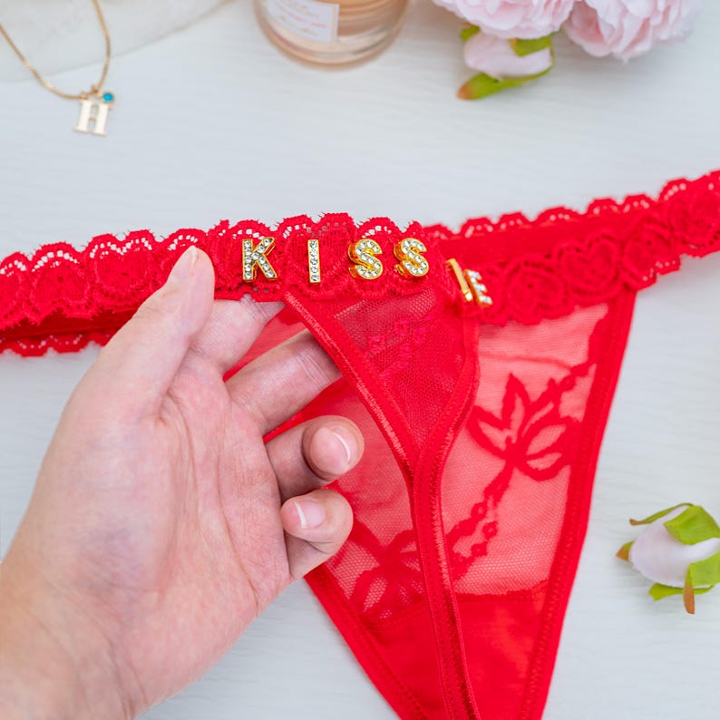 Peut inclure: String rouge avec le mot "KISS" &eacute;pel&eacute; en lettres de strass dor&eacute;es. Le sous-v&ecirc;tement a une bordure en dentelle et un motif floral sur le devant. Une main tient le sous-v&ecirc;tement.