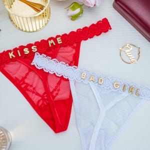 Peut inclure: Deux ensembles de lingerie. L'un est rouge avec le texte "KISS ME" en lettres dor&eacute;es. L'autre est blanc avec le texte "BAD GIRL" en lettres dor&eacute;es. Les deux ont une bordure en dentelle.