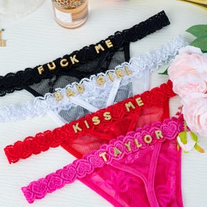 May include: Four pairs of lingerie in black, white, red, and pink. Each pair features a lace trim and gold-colored text: "FUCK ME", "BAD GIRL", "KISS ME", and "TAYLOR". The lingerie is made of sheer fabric.