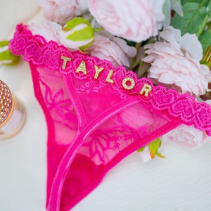 Peut inclure: String rose vif avec un motif de dentelle florale et une bordure en dentelle d&eacute;corative. Le mot "TAYLOR" est &eacute;pel&eacute; en lettres dor&eacute;es sur la ceinture. Un bouquet de fleurs roses est en arri&egrave;re-plan.