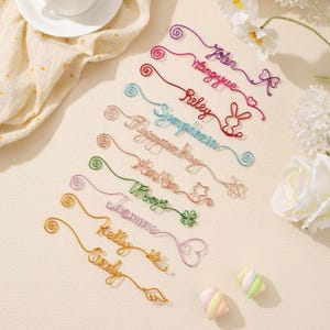 May include: A collection of colorful wire name bookmarks in various colors, including purple, red, blue, and gold. Each bookmark features a different name, such as "John" and "Riley", and decorative elements like hearts and stars. The bookmarks are arranged on a light-colored surface.