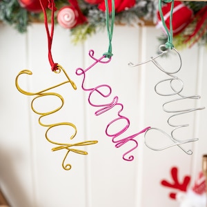 Puede incluir: Tres adornos con nombres de alambre: "Jack" en dorado, "Sarah" en rosa y "Polly" en plateado. Cada nombre está hecho de alambre y suspendido por una cinta a juego. Decoración festiva navideña con fondo blanco y adornos navideños.