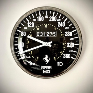Könnte beinhalten: Schwarze und silberne Ferrari F40 Tachometeruhr. Das Zifferblatt hat weiße Ziffern und Markierungen, mit einer zentralen Anzeige, die die Zahl 031275 anzeigt. Das Ferrari-Logo ist unten sichtbar.