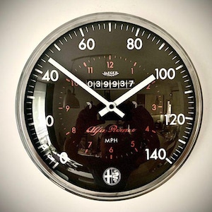 Peut inclure: Une horloge murale ronde avec un cadran noir et un bord en métal argenté. Le cadran comporte des chiffres blancs, un design de compteur de vitesse et les mots "Alfa Romeo" en rouge. Les aiguilles sont blanches.