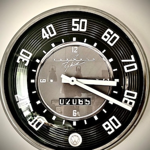 May include: A classic speedometer clock with a black and silver face. The clock has white numerals, a needle, and the text "Karmann Ghia". The odometer reads 02065. The clock is in a silver metal frame.