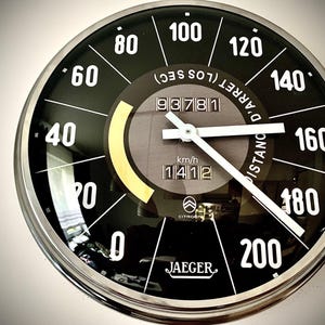 Könnte beinhalten: Eine runde schwarz-silberne Uhr im Tachometer-Design. Das Zifferblatt hat weiße Ziffern, einen weißen Zeiger und den Text "km/h" und "JAEGER". Die Uhr misst bis zu 200 km/h.