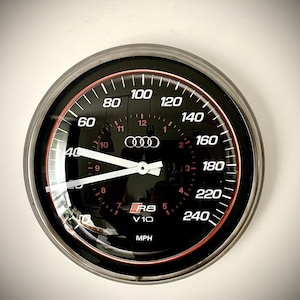 Könnte beinhalten: Ein schwarzer Tachometer mit weißen und roten Markierungen, mit dem Audi-Logo und dem Text "R8 V10". Das Messgerät zeigt Geschwindigkeiten bis zu 386 km/h an. Die Zeiger zeigen 64 km/h an.