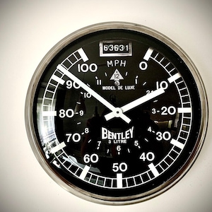 May include: A black Bentley Model de Luxe speedometer clock with white numerals and hands. The clock face displays MPH, and the text "3 Litre". The clock has a silver metal rim.