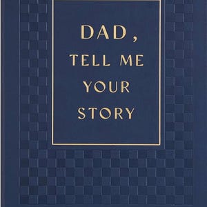 May include: A navy blue journal with a textured cover. The cover features a gold-framed rectangle with the words "DAD, TELL ME YOUR STORY" in gold lettering. A blue ribbon bookmark is visible.