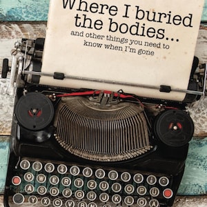 Könnte beinhalten: Eine schwarze Vintage-Schreibmaschine mit einem cremefarbenen Papier mit dem Text "Where I buried the bodies..." und anderem Text. Die Schreibmaschine hat einen Metallwagen und Tasten, vor einem verwitterten Holzgrund.