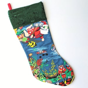 Puede incluir: Un calcetín navideño con un fondo azul que presenta a Papá Noel buceando, rodeado de coloridos corales, peces y buceadores. La parte superior del calcetín es verde y tiene un lazo rojo para colgar.