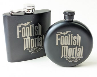 May include: Two black metal hip flasks with a silver lid. Both flasks have the same design, featuring a bat, a spider web, and the text "Foolish Mortal".