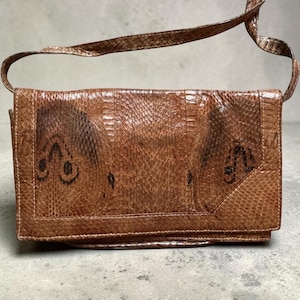 Peut inclure: Un sac à bandoulière rectangulaire marron avec un motif texturé en peau de serpent. Le sac possède une fermeture à rabat et une fine bandoulière assortie. La surface présente une texture écailleuse avec des marques marron plus foncées.