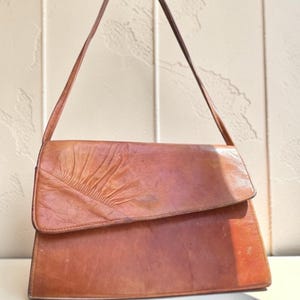Può includere: Una borsa in pelle color cuoio con una singola tracolla e chiusura a patta. La patta è decorata con una distinta trama plissettata o arricciata sul lato sinistro, creando un motivo a ventaglio sulla superficie liscia della pelle della borsa.