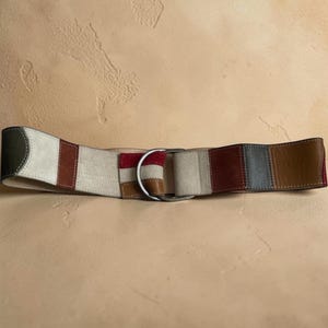 Op de afbeelding: Een brede riem met een patchworkontwerp van suède en leren panelen in de kleuren crème, bordeaux, bruin en donkergrijs. De riem wordt vastgemaakt met een zilverkleurige metalen dubbele D-ring gesp en is voorzien van zichtbare stiksels langs de randen.