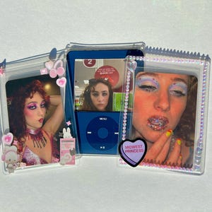 Puede incluir: Una funda de pl&aacute;stico transparente con tres fotos y un iPod. Las fotos muestran a una persona maquillada. El iPod es azul. La funda tiene dijes de coraz&oacute;n y conejito rosas. Una foto tiene un coraz&oacute;n que dice "Hot to go". Otra tiene un coraz&oacute;n que dice "Midwest Princess".