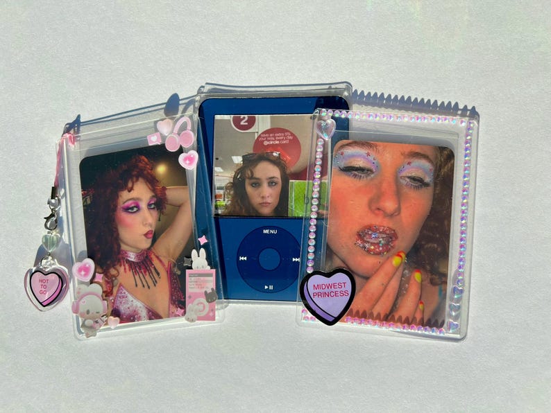 Puede incluir: Una funda de pl&aacute;stico transparente con tres fotos y un iPod. Las fotos muestran a una persona maquillada. El iPod es azul. La funda tiene dijes de coraz&oacute;n y conejito rosas. Una foto tiene un coraz&oacute;n que dice "Hot to go". Otra tiene un coraz&oacute;n que dice "Midwest Princess".