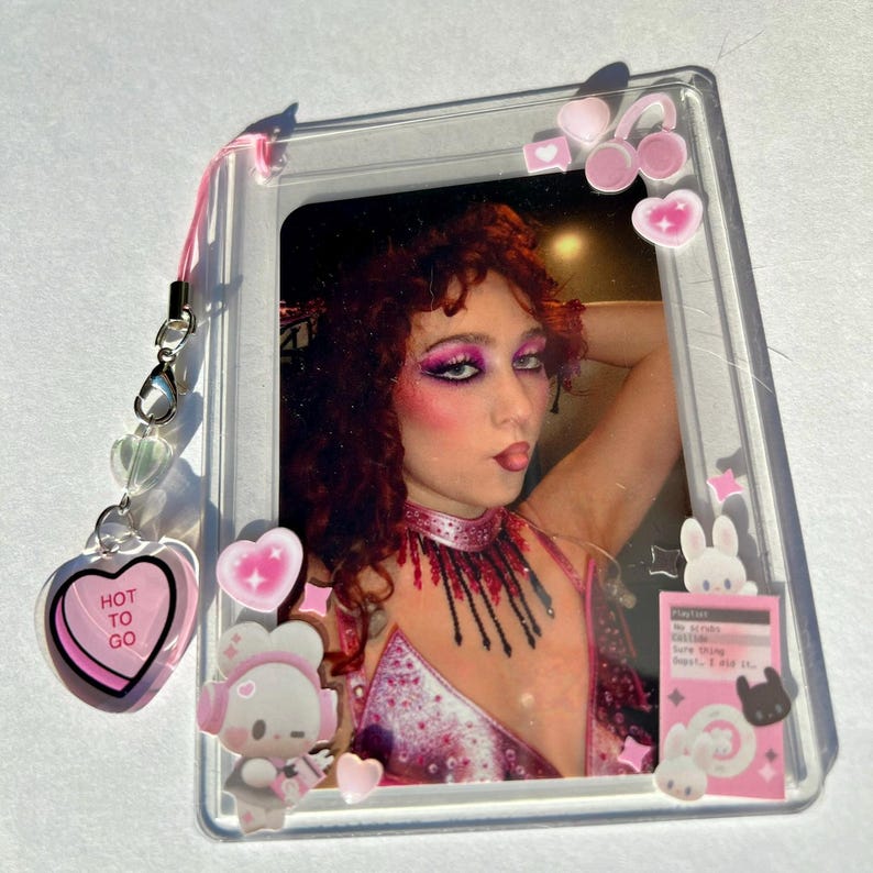 Puede incluir: Un marco de fotos de acr&iacute;lico transparente con una foto de una persona con maquillaje rosa y cabello rojo. El marco est&aacute; decorado con pegatinas rosas de coraz&oacute;n y conejito. Un dije en forma de coraz&oacute;n rosa con el texto "HOT TO GO" cuelga.