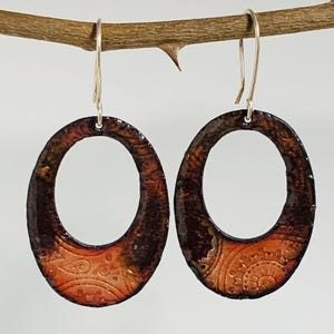 May include: A pair of oval earrings with a textured, earthy design. The earrings have a dark brown outer edge that fades into a lighter orange-brown centre with a detailed pattern. They hang from silver-coloured hooks.