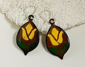 Può includere: Un paio di orecchini a forma di foglia con sfondo marrone e un disegno di tulipano giallo. Il tulipano ha foglie verdi. Gli orecchini hanno ganci color argento.