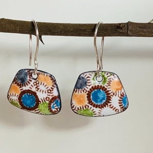 Peut inclure: Une paire de boucles d'oreilles pendantes en argent avec émail blanc et un motif géométrique coloré. Le motif présente des couleurs bleues, orange, brunes et vertes.