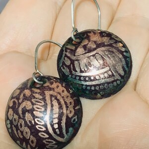 Peut inclure: Une paire de boucles d'oreilles pendantes en argent avec des disques ronds de couleur violet foncé et cuivre. Les disques présentent un motif paisley argenté.