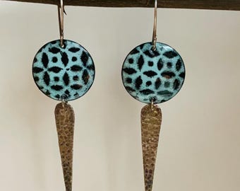May include: A pair of handcrafted earrings. Each earring features a round, turquoise-colored enamel top with a black geometric pattern, connected to a textured, bronze-colored, elongated teardrop shape. The earrings hang from silver hooks.