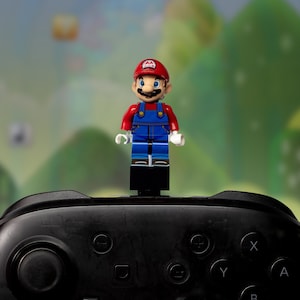 May include: A black video game controller with a red and blue Lego figure of Mario standing on top of it. The background is a blurred image of a green and brown landscape.